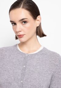 Cardigan grigio lavorato a maglia con scollo rotondo, rifinitura bianca e chiusura con bottoni. Tessuto morbido con un motivo discreto su tutta la superficie.