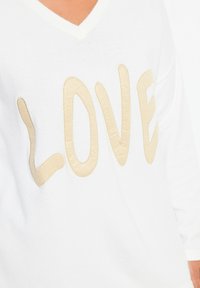 Maglione bianco con scollo a V, caratterizzato da un grande testo in rilievo beige "LOVE", texture liscia e maniche lunghe.