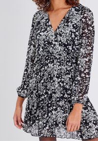 Robe noire à motifs floraux avec de longues manches transparentes. Elle présente un décolleté en V, une taille froncée et une jupe plissée. Motif floral dans des tons de blanc.