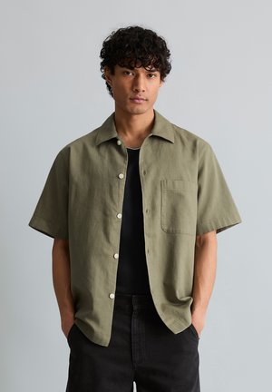 Junger Mann mit lockigem Haar, der ein offenes olivgrünes kurzärmeliges Hemd über einem schwarzen T-Shirt trägt, die Hände in den Taschen.