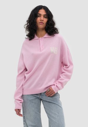 Kvinde med krøllet mørkt hår iført en løs lyserød sweater med krave og let logo på brystet samt lyseblå jeans, hånd på hoften.