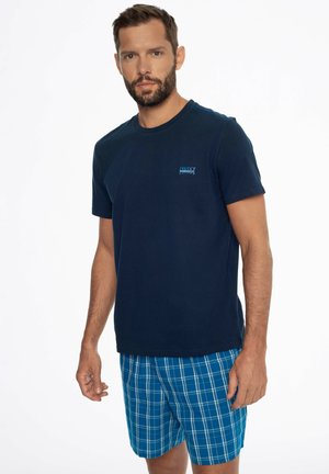 Mann trägt dunkelblaues kurzärmeliges T-Shirt und blaue karierte Shorts und steht vor weißem Hintergrund.