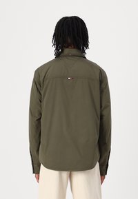 Olivengrøn letvægtsjakke med knappet krave, rygstykke-detalje og logoaccents. Lange ærmer med knapper ved manchetterne, glat tekstur.