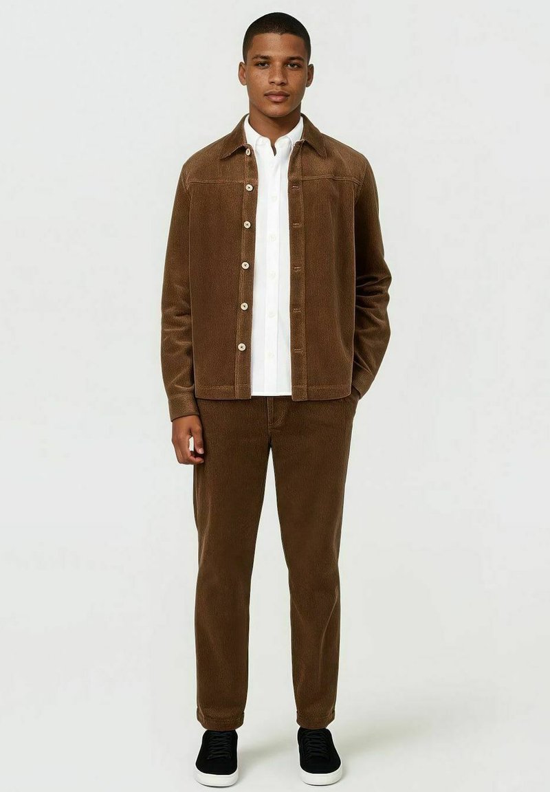 Brown corduroy jas en bijpassende broek, met een wit shirt eronder. De jas heeft een knoopsluiting en een zachte textuur.