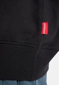 Zwarte sweatshirt met een gestructureerd breipatroon, met een rood label aan de linkerkant nabij de zoom. Samengevoegd met blauwe stof eronder.