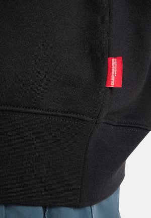 Sweat-shirt noir avec un motif en tricot texturé, orné d'une étiquette rouge sur le côté gauche près de l'ourlet. Fusionne avec un tissu bleu en dessous.