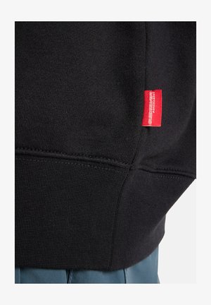 Black sweatshirt featuring a textured knit design, with a red label on the left side near the hem. Merges with blue fabric underneath.