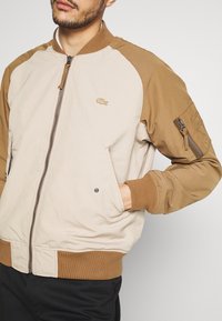 Homme portant une veste bomber beige et marron avec fermeture éclair à l'avant et poche zippée sur la manche, main dans la poche avant, pantalon noir visible.