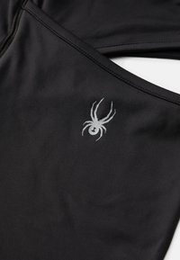 Sorte atletiske bukser med en glat tekstur, prydet med et sølvfarvet edderkoppe-logo på den nederste del og kontrasterende syninger.