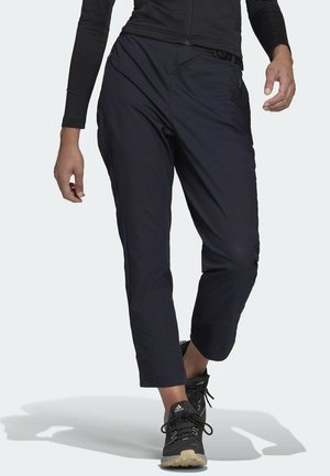 Persoon loopt en draagt zwarte taps toelopende broek, zwarte jas met rits en zwarte Adidas-sneakers met beige zolen.