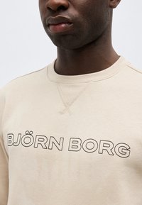 Κοντινό πλάνο ενός ατόμου που φοράει μπεζ φούτερ με την επιγραφή "BJÖRN BORG" σε μαύρα γράμματα στο στήθος.