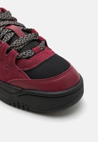 Sneaker rosso e nero con una combinazione di materiali in suede e mesh, superficie testurizzata, punta tondeggiante e lacci grigi in contrasto.