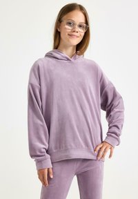 Lavenderfarbene Kapuzenjacke aus weichem, plüschigem Stoff mit einer großen Kapuze, überschnittenen Schultern und gerippten Bündchen. Kombiniert mit passender Unterbekleidung.