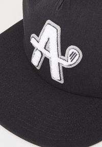 Black cap made of fabric featuring a prominent white embroidered letter "A" with a textured accent. Flat brim and detailed stitching visible.