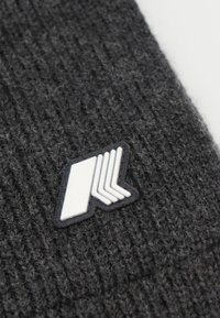 Cappello in maglia grigio con superficie testurizzata, caratterizzato da una patch in gomma bianca con logo su sfondo nero, posizionata sul lato.
