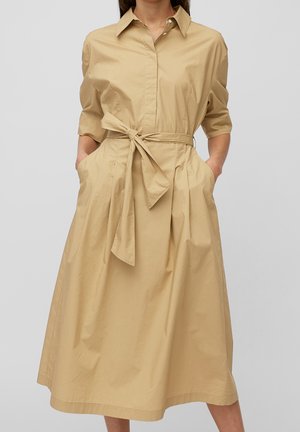 Vrouw draagt beige midi overhemddress met halve mouwen, kraag, knopen aan de voorkant, zijzakken en een stoffen riem gestrikt in de taille.