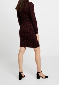 Donna in piedi di spalle, indossa un abito aderente a maniche lunghe, lungo fino al ginocchio, con stampa leopardata nera e rosso scuro e scarpe con tacco a blocco nere.