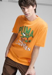Oranžs kokvilnas T-krekls ar karikatūras grafiku, kas attēlo zaļu antropomorfu tēlu, ar tekstu "PUMA Carrots" zaļā un baltā krāsā.