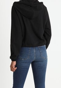 Sudadera negra recortada con capucha y dobladillo elástico, combinada con jeans ajustados azules que cuentan con bolsillos traseros y detalles de costura sutiles.