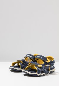 Morske plave i žute dječje sandale s podesivim čičak trakama, otvorenim prstima i uzorkovanim gumiranim potplatom. Uključuje Timberland logotip.