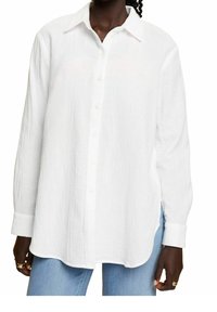 Chemise blanche à manches longues, à boutons, en tissu texturé. Elle présente un col classique, des boutons à l'avant et des fentes sur les côtés au niveau de l'ourlet.