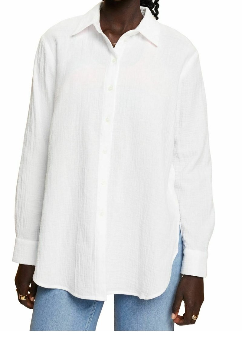 Chemise blanche à manches longues, à boutons, en tissu texturé. Elle présente un col classique, des boutons à l'avant et des fentes sur les côtés au niveau de l'ourlet.