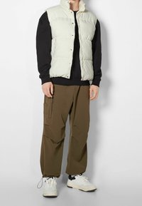 Fjäderlätt ljusgrön väst med hög krage, svart sweatshirt under, olivgröna cargobyxor och vita sneakers med svarta detaljer.