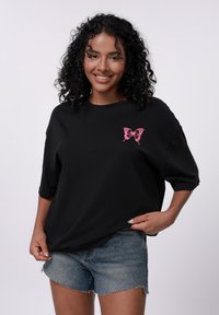 Svart oversized t-skjorte med en rosa sommerfugl grafikk på venstre bryst, kombinert med denimshorts. Glatt stoff, avslappet passform.