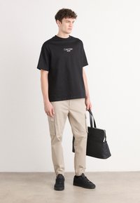 Zwarte katoenen T-shirt met het logo van "Calvin Klein Jeans", beige cargobroek met zijzakken, zwarte sneakers en een zwarte totebag.