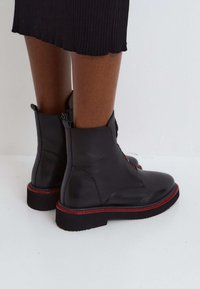 Bottes de cheville en cuir noir avec une finition lisse, accent en couture rouge et une semelle à plateforme basse. Fermeture éclair sur le côté.