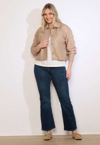 Lachende vrouw met een beige cropped jack, wit topje, blauwe uitlopende jeans en beige mocassins, staand tegen een witte muur.