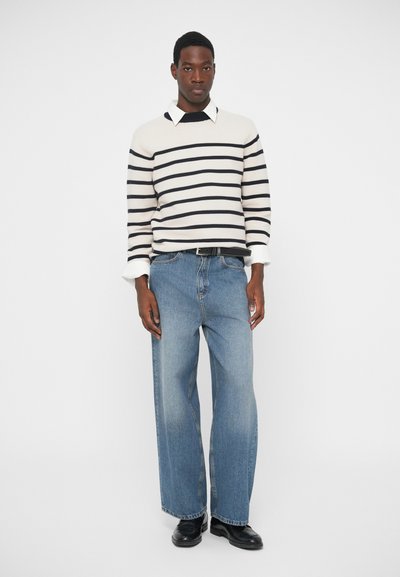 Prugasta puloverica u krem i tamnoplavoj boji, uparena s ležernim plavim trapericama. Odjeća ima ovratnik i crni pojas; crna obuća upotpunjuje izgled.