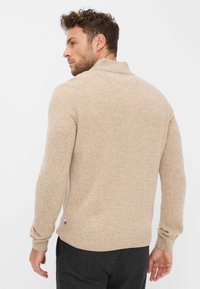 Beiger Strickpullover mit hohem Kragen, gerippten Bündchen und Saum. Strukturierter Stoff, lockere Passform und ein kleines Logodetail an der Seite.
