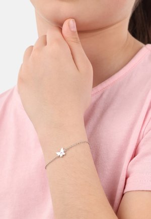 Silberne Kettenarmband mit einem Schmetterlingsanhänger und einem kleinen Edelsteindetail, getragen an einer Hand gegen ein pinkes Hemd.