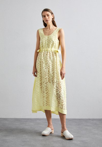 Robe midi en dentelle jaune avec un décolleté en scoop et une taille à nouer. Présente un design semi-transparent et est associée à des sabots blancs.