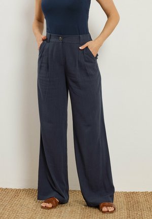 Frau trägt dunkelblaue, hoch taillierte, weit geschnittene Hose mit vorderen Falten und Seitentaschen, braune Sandalen mit offenem Zehenbereich und ein ärmelloses, marineblaues Oberteil.