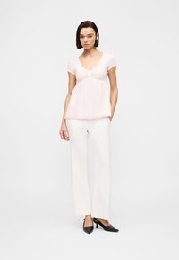 Γυναίκα που στέκεται με κοντά σκούρα μαλλιά, φοράει ροζ ανοιχτό ροζ μπλούζα με κοντά μανίκια, λευκό παντελόνι με φαρδιά πόδια και μαύρα παπούτσια με τακούνι.