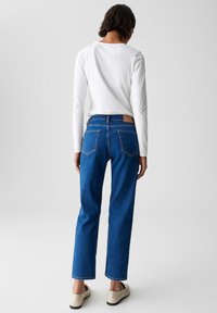 Personne de dos, portant une chemise blanche à manches longues, un jean bleu droite et des chaussures beiges à enfiler, sur un fond gris uni.