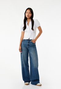 Witte kortemouwen overhemd met pofmouwen, gecombineerd met oversized blauwe denim jeans. Model draagt beige sneakers.