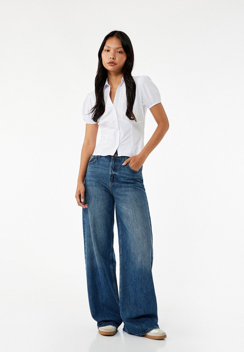 Witte kortemouwen overhemd met pofmouwen, gecombineerd met oversized blauwe denim jeans. Model draagt beige sneakers.