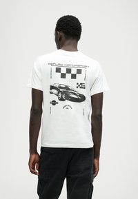 Biała bawełniana koszulka z czarną grafiką samochodu na plecach, z napisem "REPLAY MOTORSPORT" i szachownicowymi akcentami dookoła.