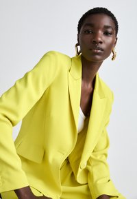 Bright yellow blazer with a tailored fit, single button closure, sharp lapels, and smooth texture, paired with a white top.