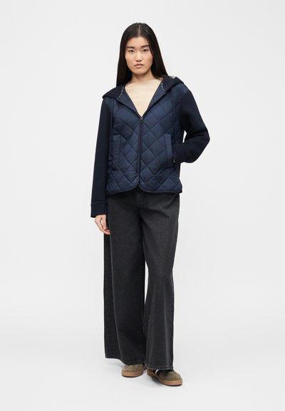 Jeune femme portant une veste matelassée bleu marine zippée avec capuche, un haut beige, un jean noir à jambes larges, et des chaussures marron à enfiler, debout avec une main dans la poche.