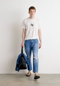 Camiseta blanca con un pequeño gráfico, jeans azules desgastados, sandalias negras, llevando una bolsa de mano negra y una chaqueta de mezclilla colgada del brazo.