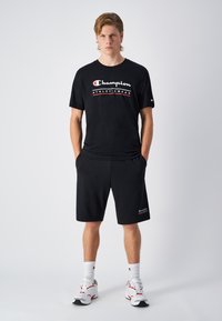 Zwarte sportt-shirt met het witte en rode logo "Champion ATHLETICWEAR", gecombineerd met bijpassende zwarte shorts en witte sneakers.