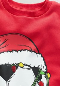 Sweatshirt roșu cu un grafic al feței lui Moș Crăciun, împodobit cu o pălărie cu margine din blană albă și accente de lumini colorate de sărbătoare.