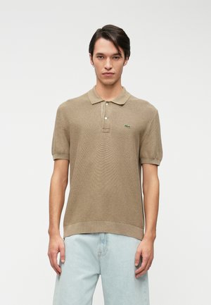 Camisa polo beige de manga corta, con cuello, tapeta de tres botones y logo bordado. Lleva puesta con pantalones vaqueros azul claro. Textura suave.