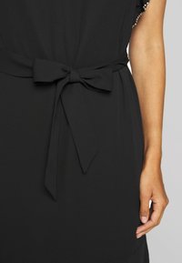 Robe noire avec ceinture en tissu nouée en un nœud à la taille et un bras visible reposant le long du corps sur un fond uni.