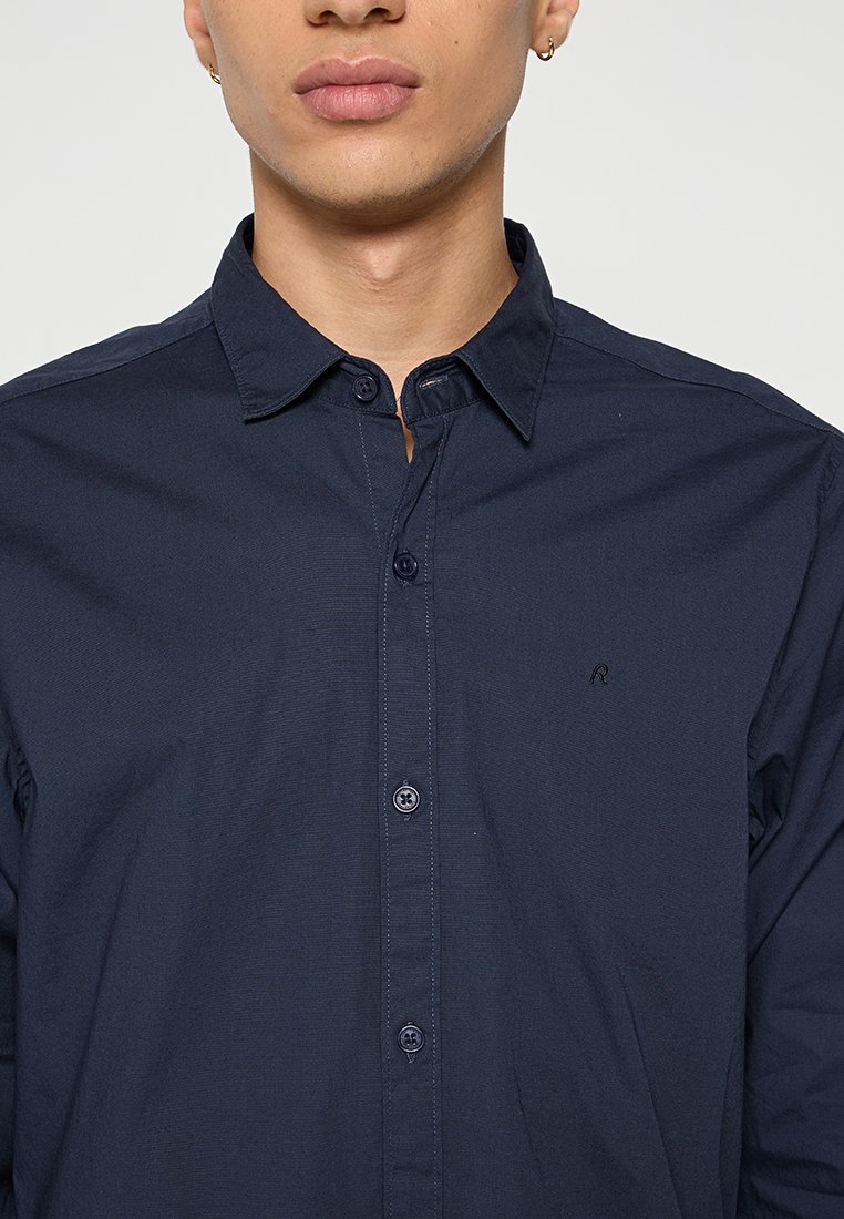 Chemise boutonnée bleu marine en coton. Elle présente un col classique, une texture subtile et un petit logo brodé sur la poitrine gauche.