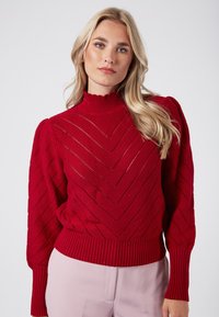 Maglione rosso lavorato a maglia con colletto alto a coste, motivo a zigzag e maniche a palloncino. Orlo e polsini a coste, realizzato in un tessuto testurizzato.